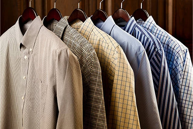 Six collared dress shirts with various checkered and striped patterns hang on wooden hangers against a wooden background.
