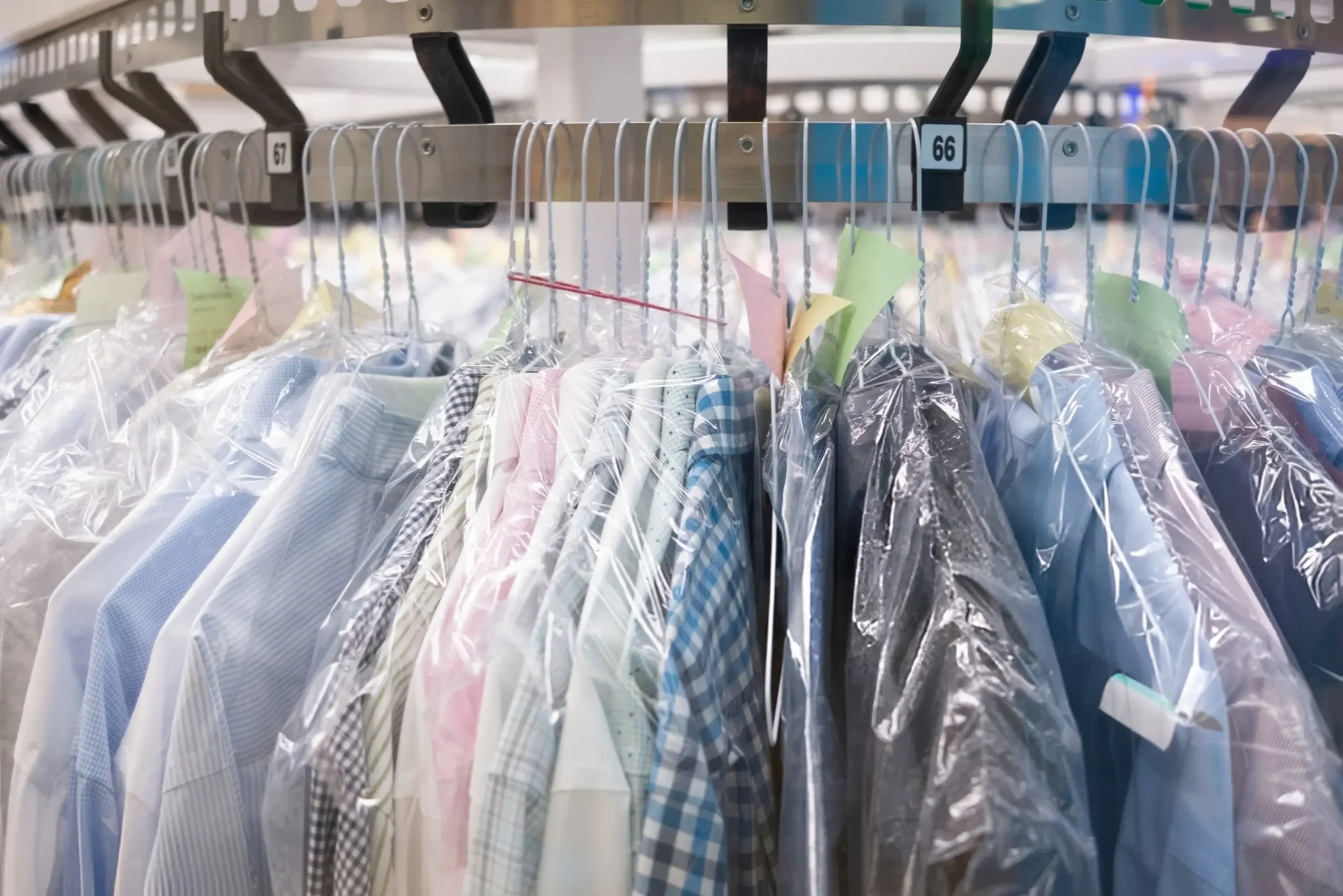 Clothes on wire hangers covered in plastic, hanging on a metal rack at a dry cleaning facility, with numbered tags visible above the garments.