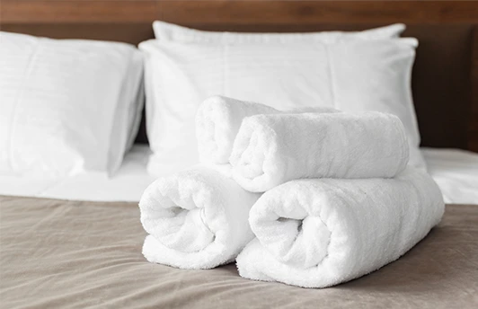 Three rolled white towels are stacked on a neatly made bed with white pillows and beige bedding.