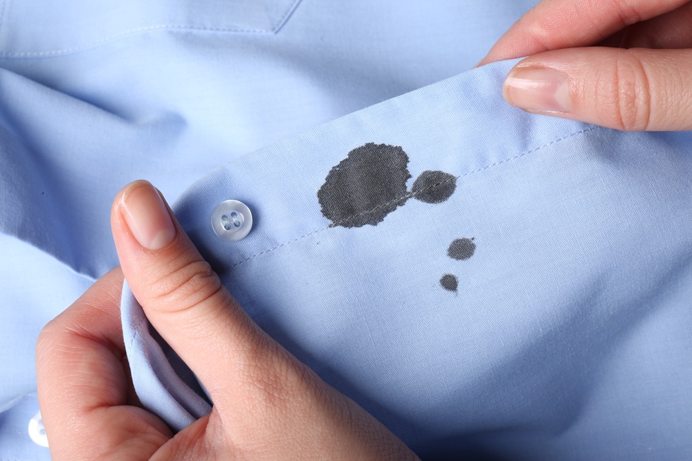 A close-up of hands holding a light blue shirt with several black ink stains on the fabric near a button.