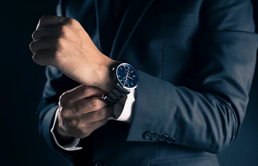 A person in a dark suit adjusts the cuff of their sleeve while wearing a wristwatch with a metal band and dark face.