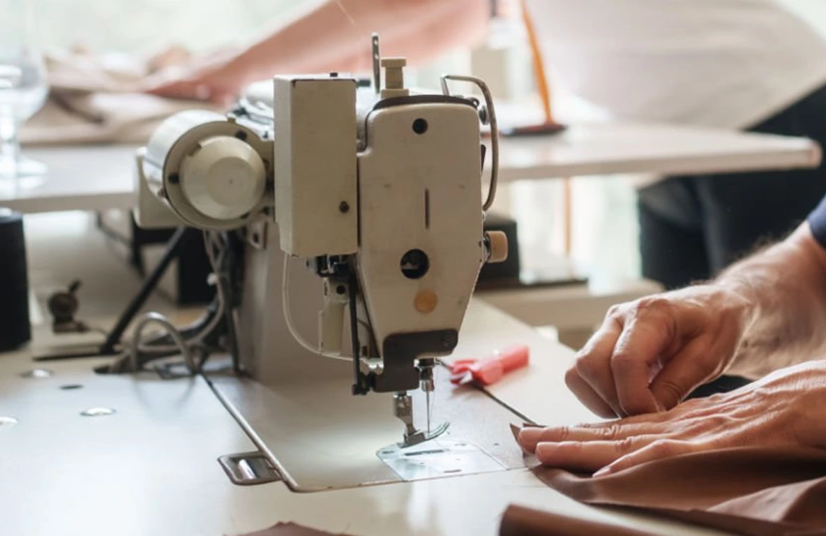 Close-up of hands guiding fabric through a sewing machine on a work table, with tools and another person visible in the background.