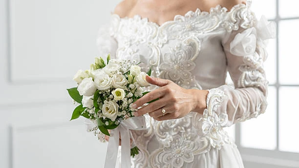 A bride in an off-the-shoulder white wedding dress holds a bouquet of white flowers and greenery, showing a ring on her left hand.