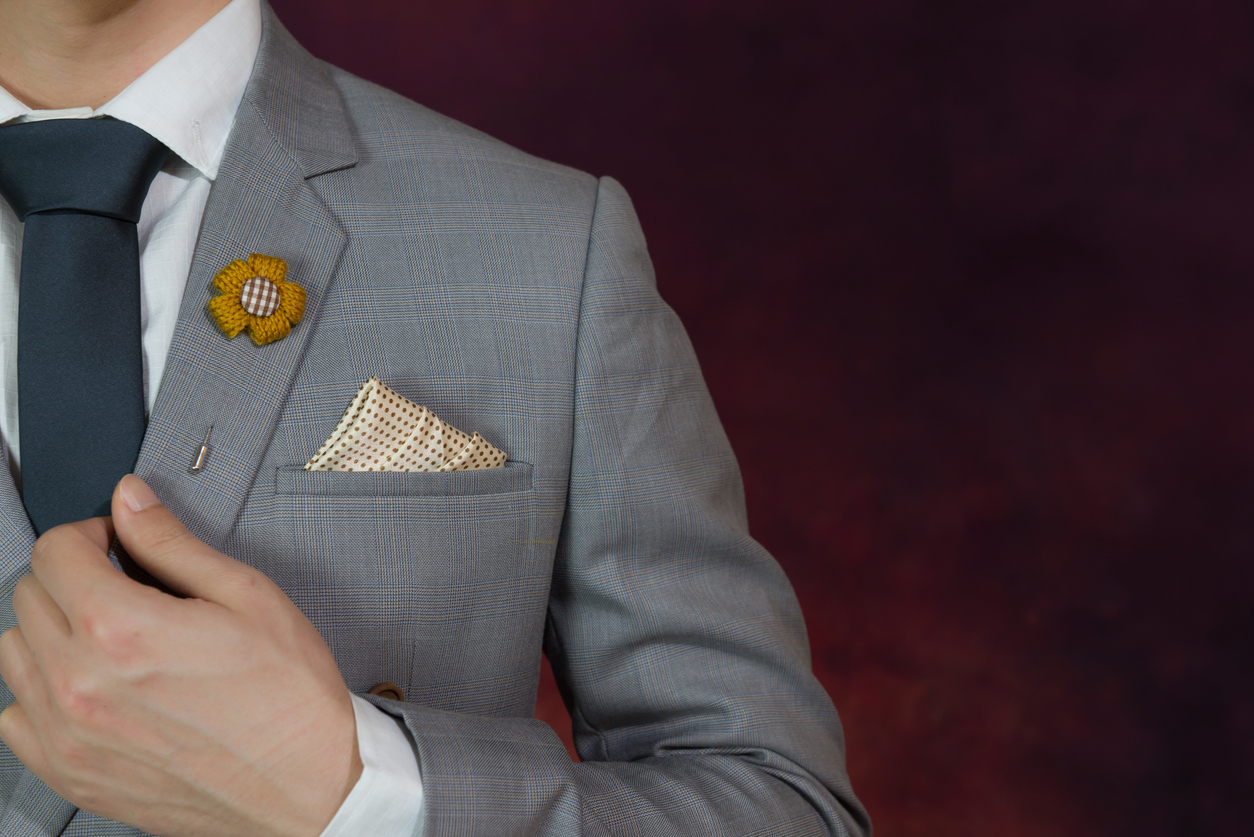 Person in a light gray suit and black tie adjusts jacket, with a yellow flower lapel pin and polka dot pocket square, against a dark, blurred background.