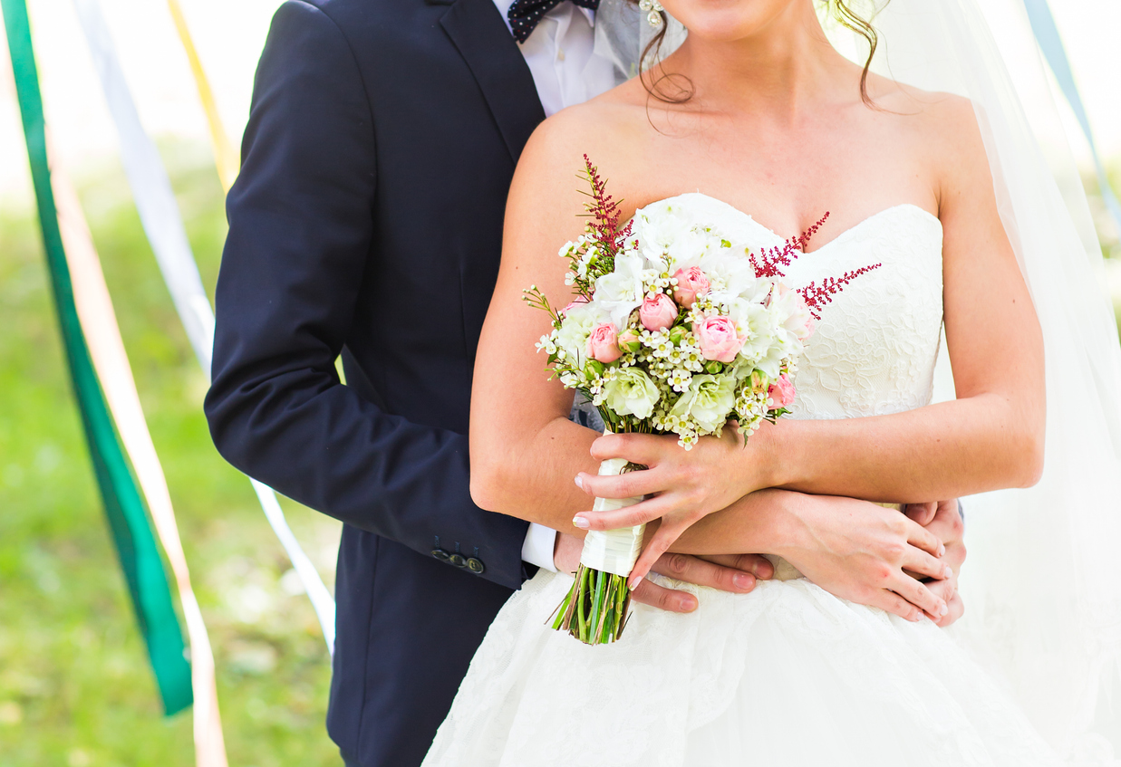 A bride in a white dress holding a bouquet is embraced from behind by a groom in a dark suit outdoors.
