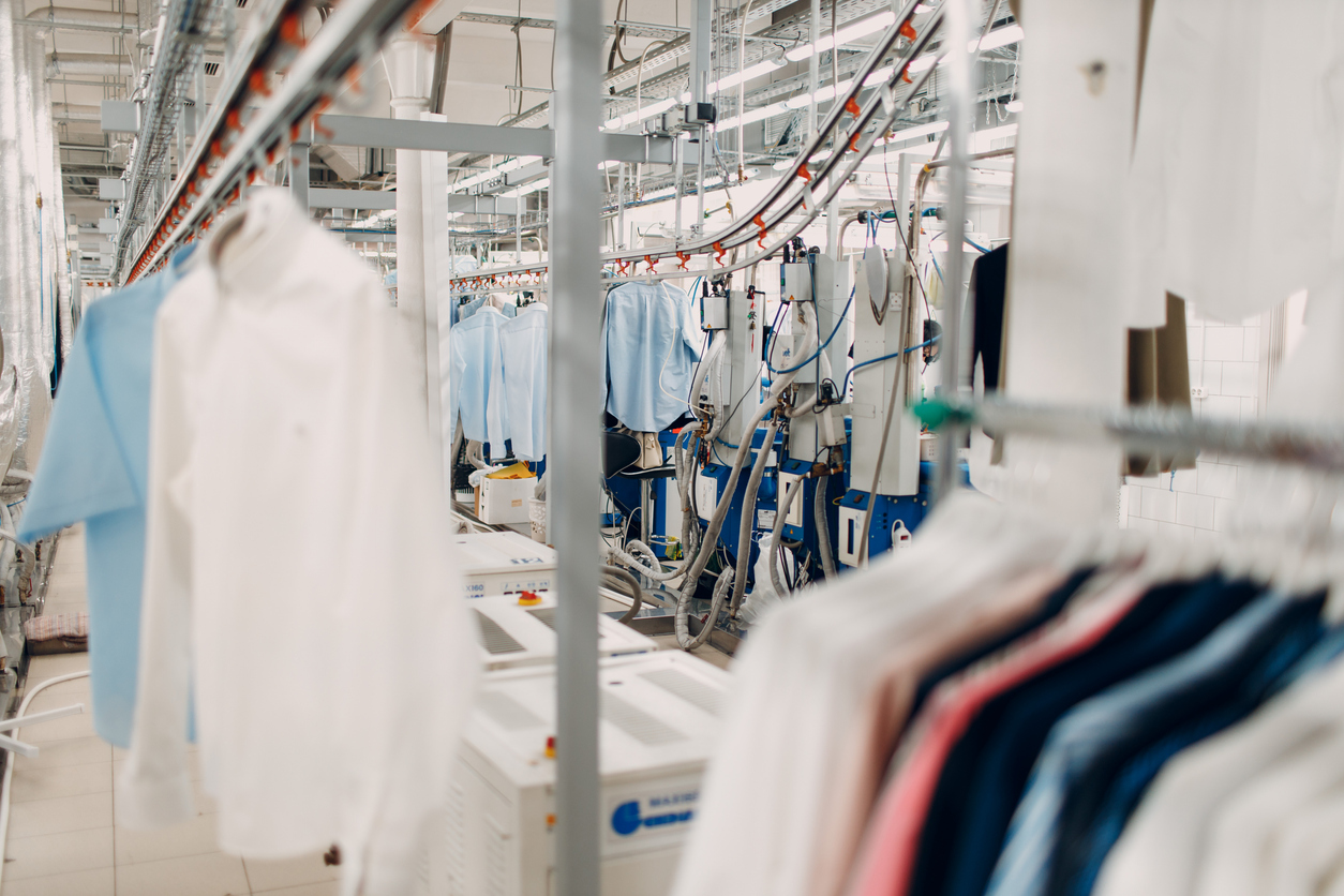 A commercial laundry facility with shirts hanging on racks and industrial washing and pressing machines in a bright, clean environment.