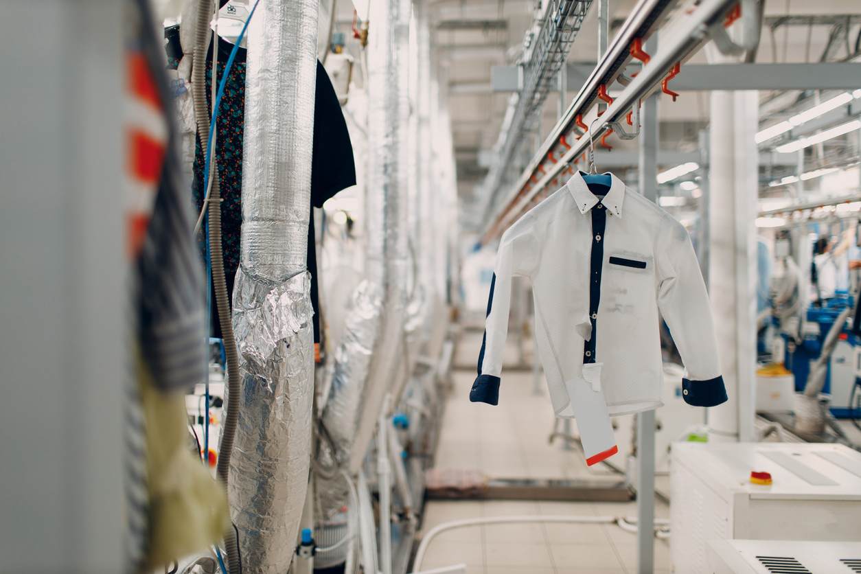 A white shirt with dark accents hangs on a conveyer in a modern garment factory, with industrial equipment and ducts visible in the background.