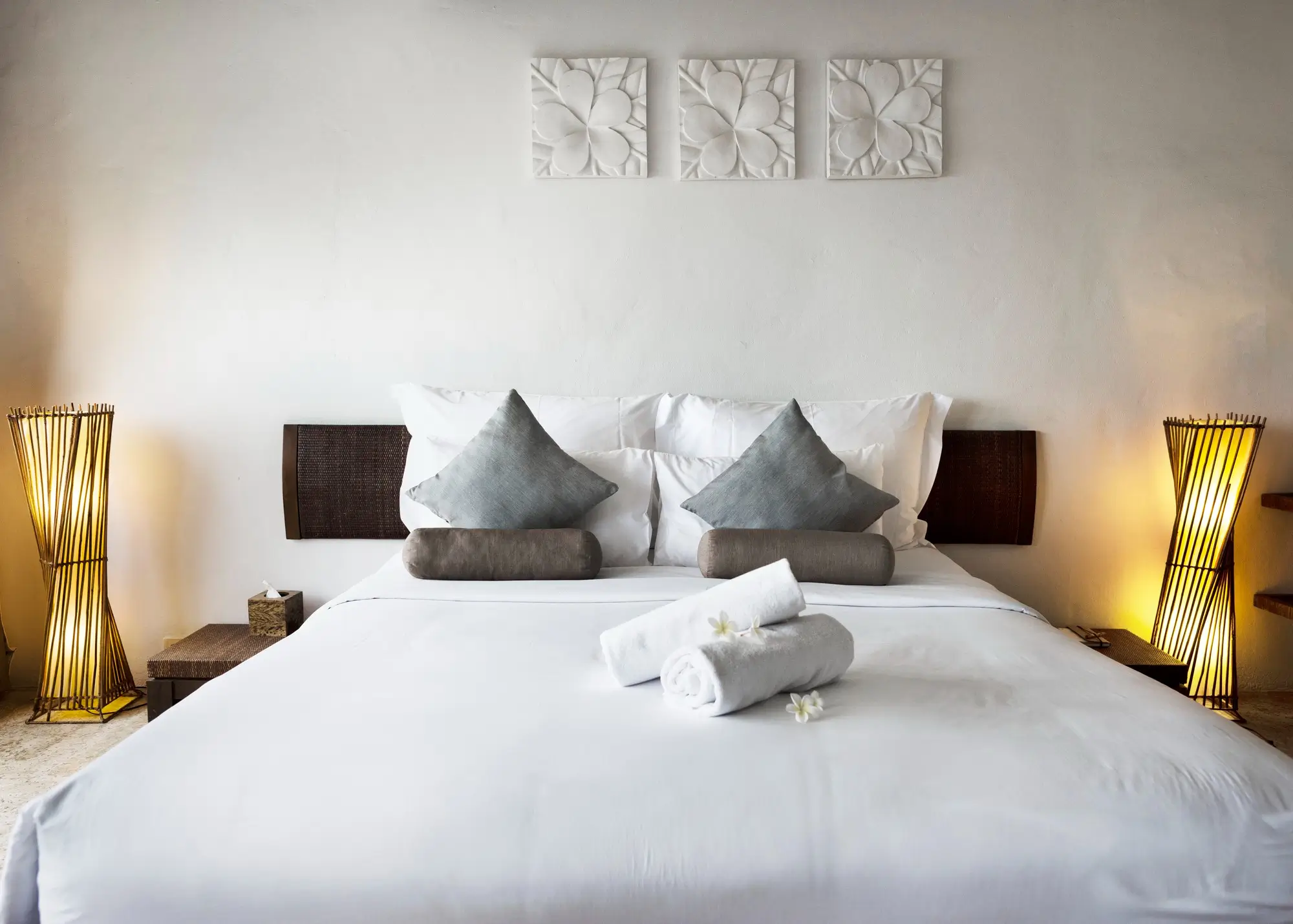 Neatly made bed with white linens, two blue-gray pillows, rolled towels, and soft lighting from two lamps, against a white wall with three decorative panels above the headboard.