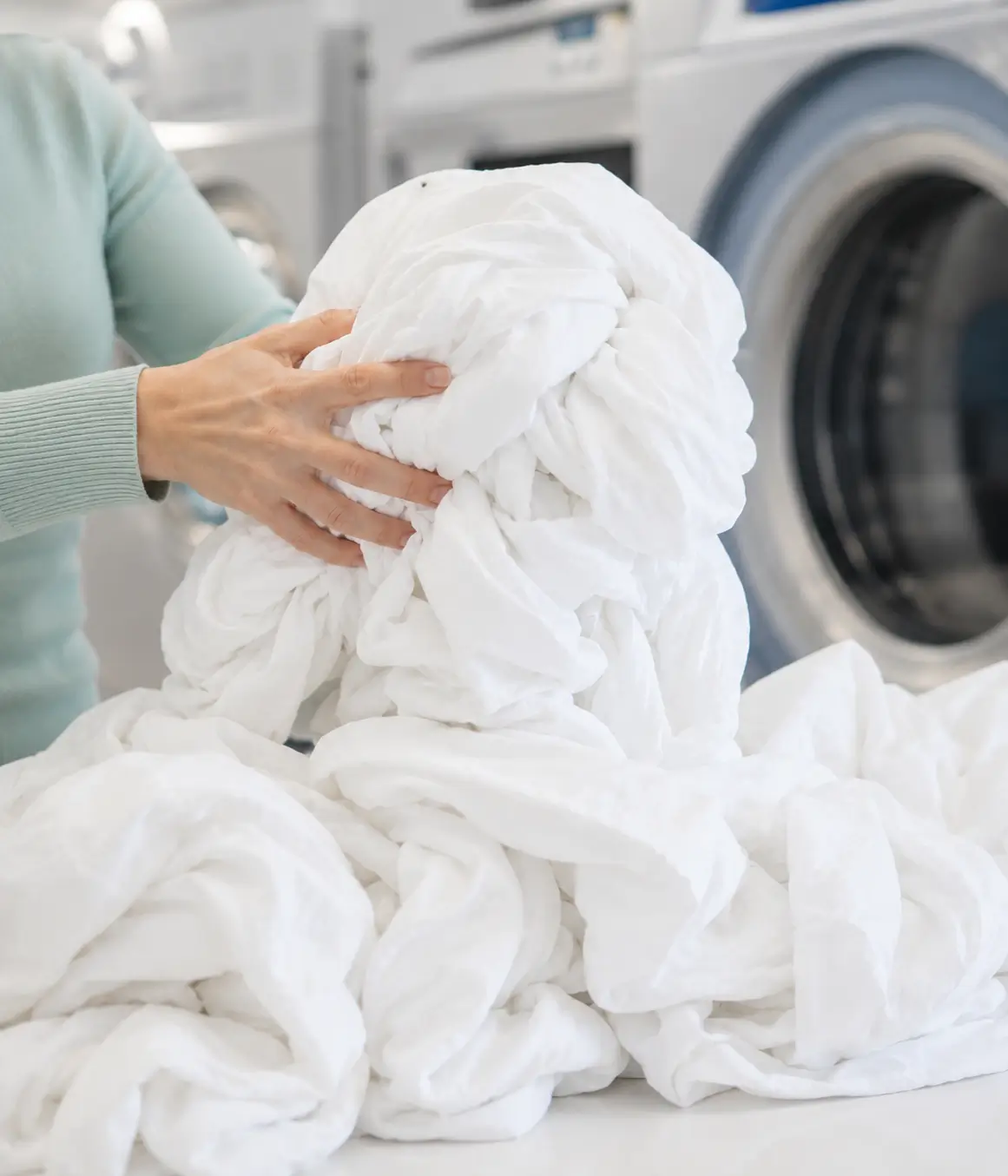 Person holding a large bundle of clean white laundry in front of industrial washing machines at a laundromat.