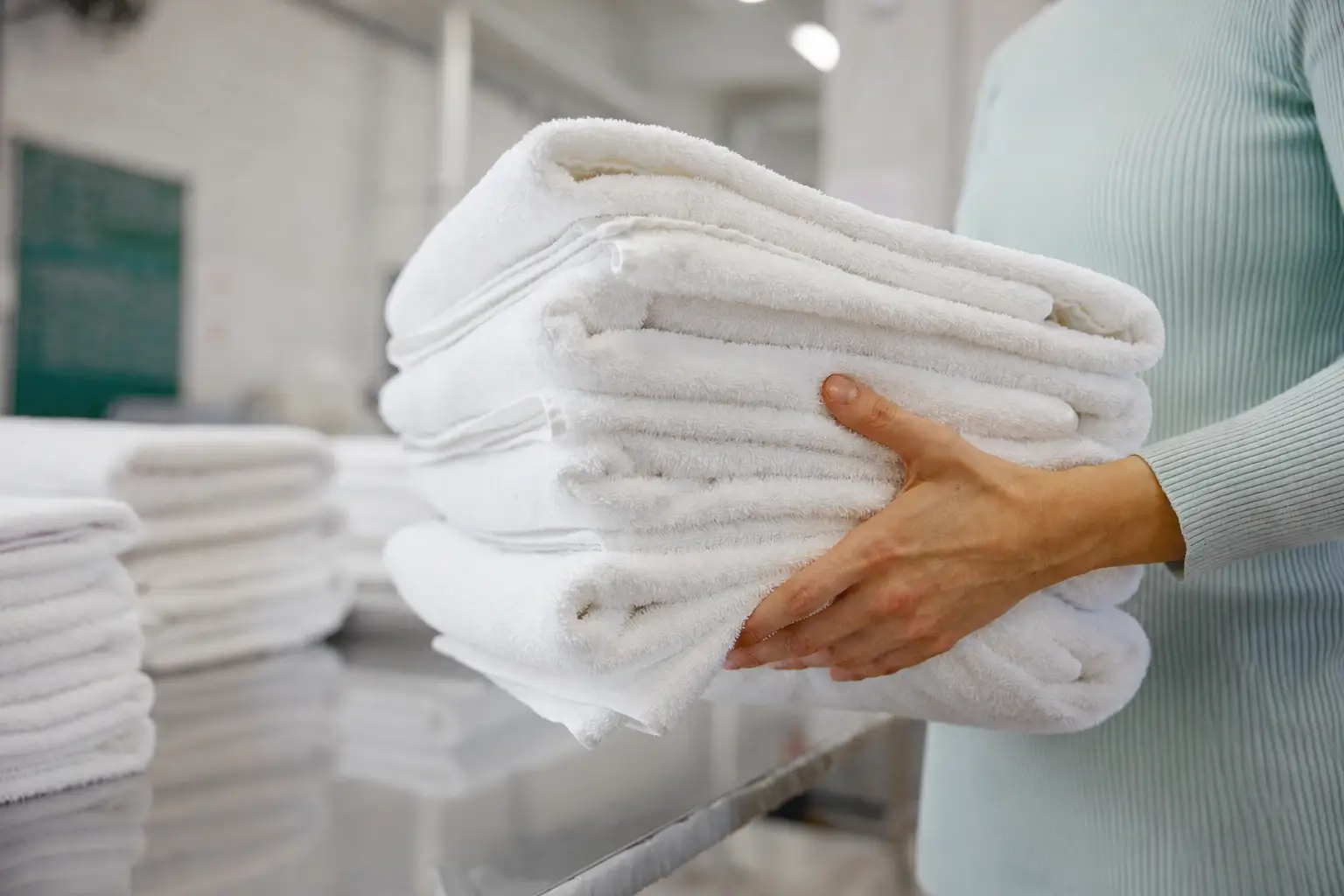 A person in a light green shirt holds a stack of folded white towels in a clean, industrial laundry setting.