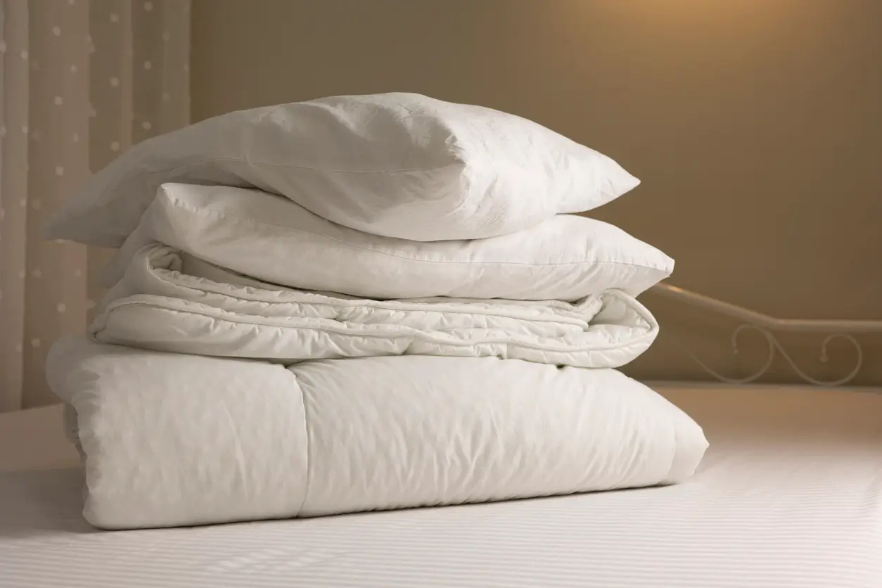 A stack of white bedding, including pillows, a comforter, and a folded quilt, sits on a neatly made bed against a neutral background.