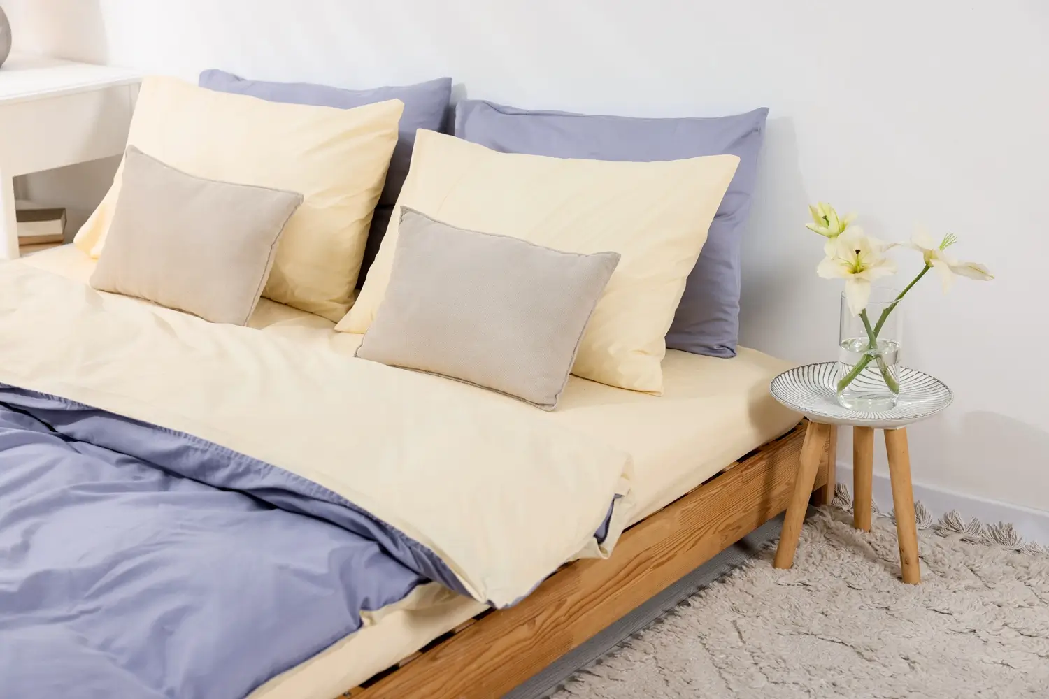 A neatly made bed with cream and lavender bedding, two large pillows, two smaller gray pillows, and a small table holding a vase with white flowers.