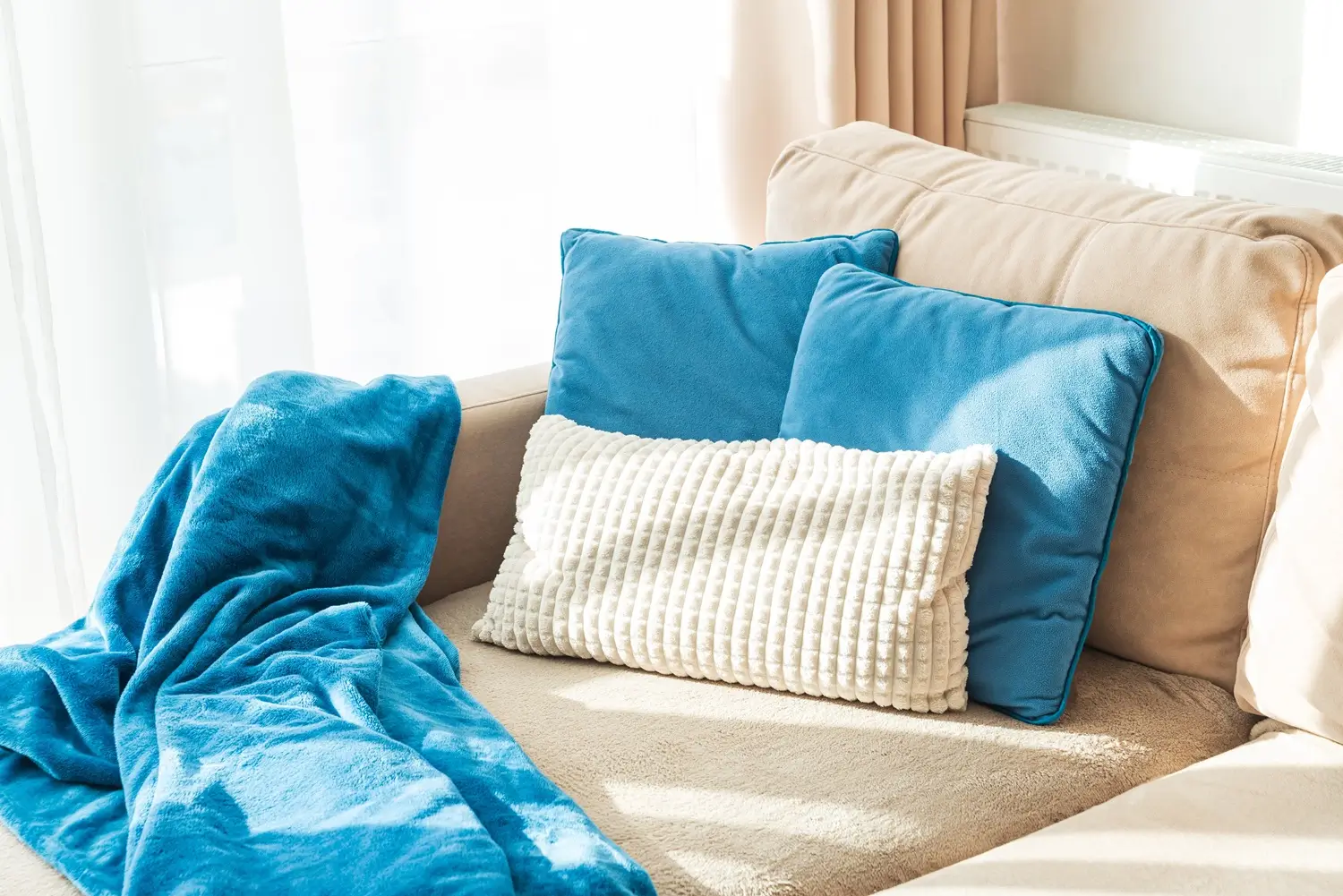 Beige sofa with two blue cushions, a textured white rectangular pillow, and a blue blanket draped over the seat, near a sunlit window with sheer curtains.