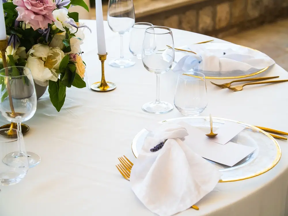 Elegant round table set with white linens, gold cutlery, clear glassware, white plates, cloth napkins, candles, and a floral centerpiece.