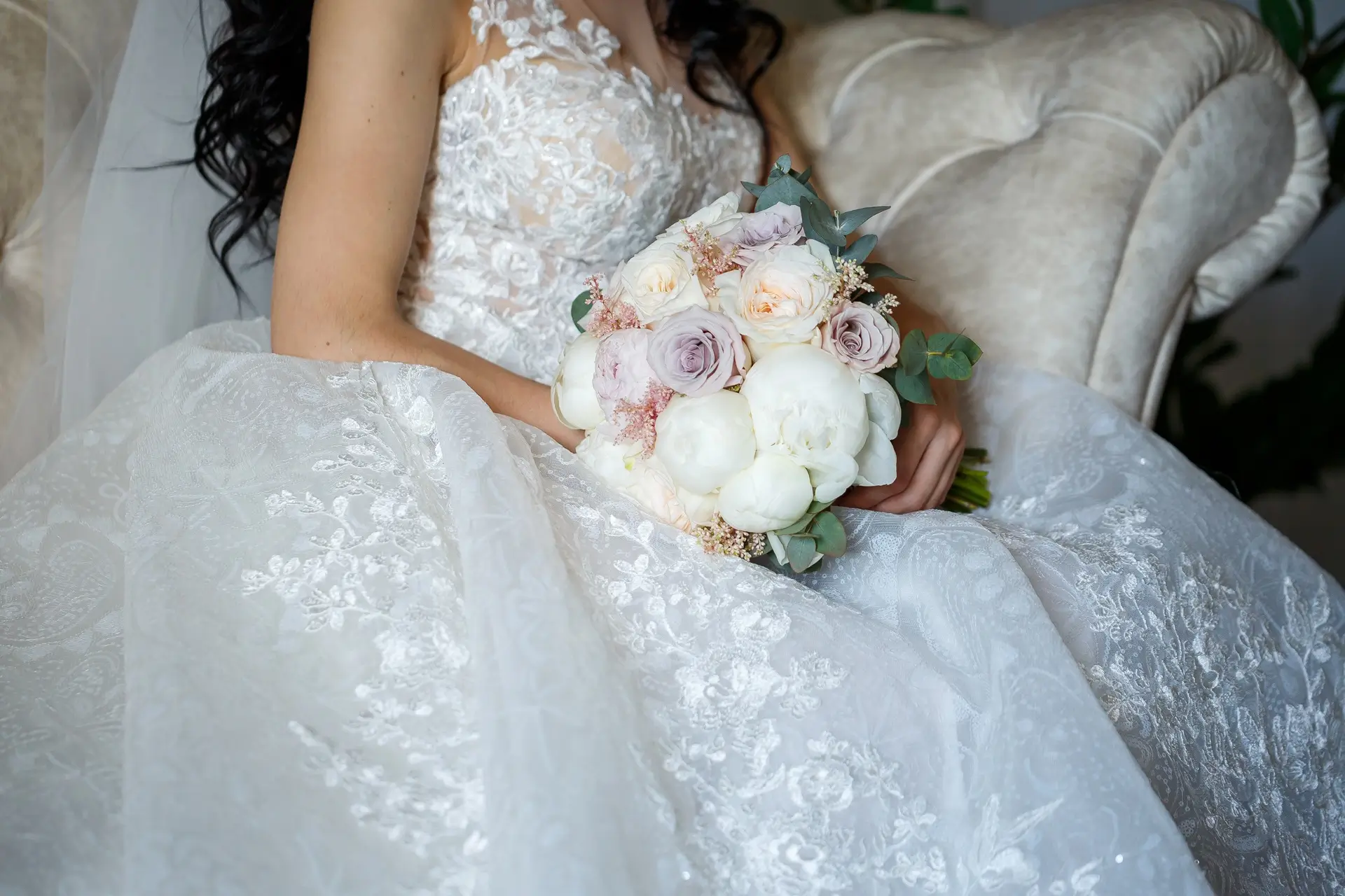 A bride in a white lace wedding dress holds a bouquet of white and pale pink roses while sitting on a cream-colored sofa.