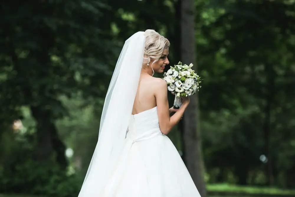 Bride in a white strapless wedding dress and veil stands outdoors, holding a bouquet of flowers, with green trees in the background.