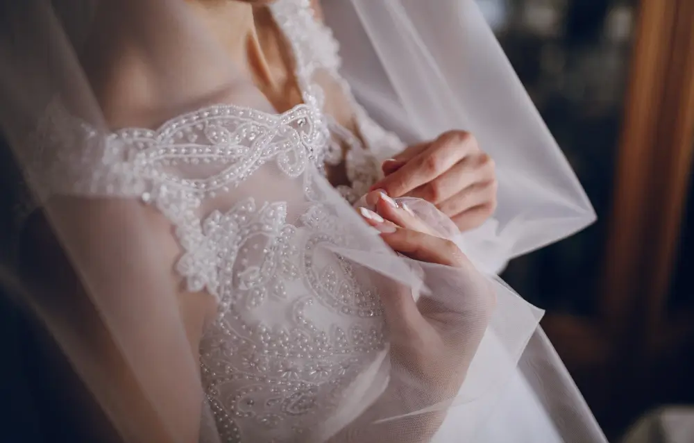 Close-up of a bride adjusting her sheer lace wedding dress with beaded details, partially covered by a translucent veil.